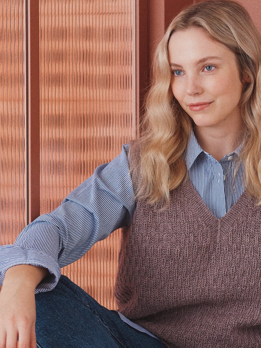 Una chica sentada, de fondo una pared naranja. La chica lleva unos vaqueros, una camisa de rayas y, sobre esta, un chaleco tejido con una textura como de trigo en un color malva apagado.